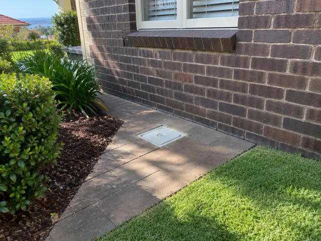 Flush inspection cover set in paved path beside brick house and lawn in Dover Heights